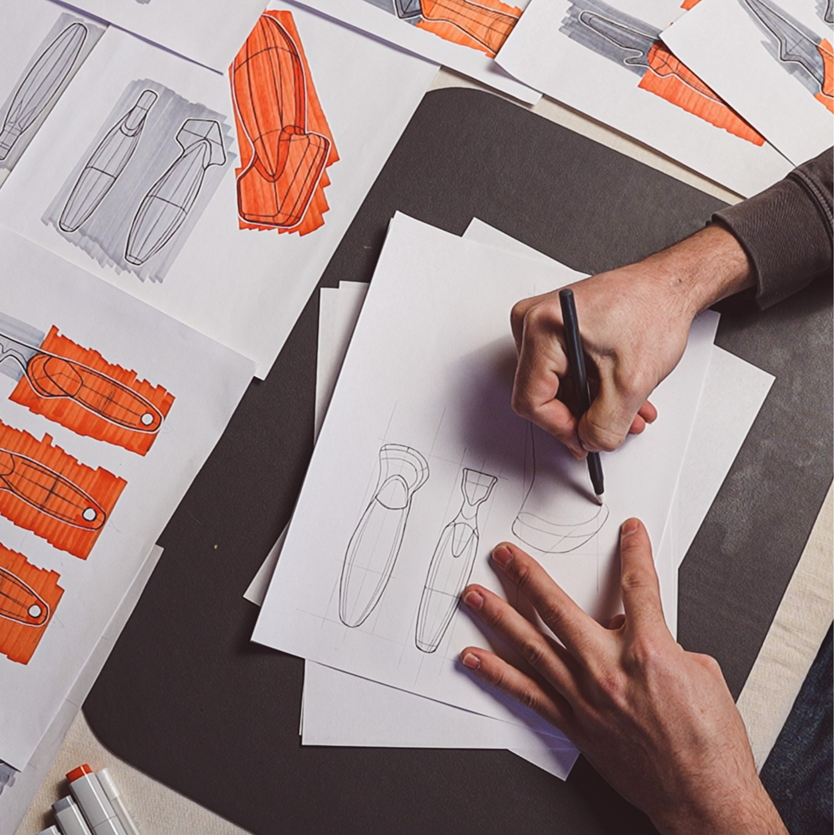 A designer’s hands sketching multiple oyster knife handle concepts on paper, surrounded by marker renderings and early shape explorations in orange and grey.