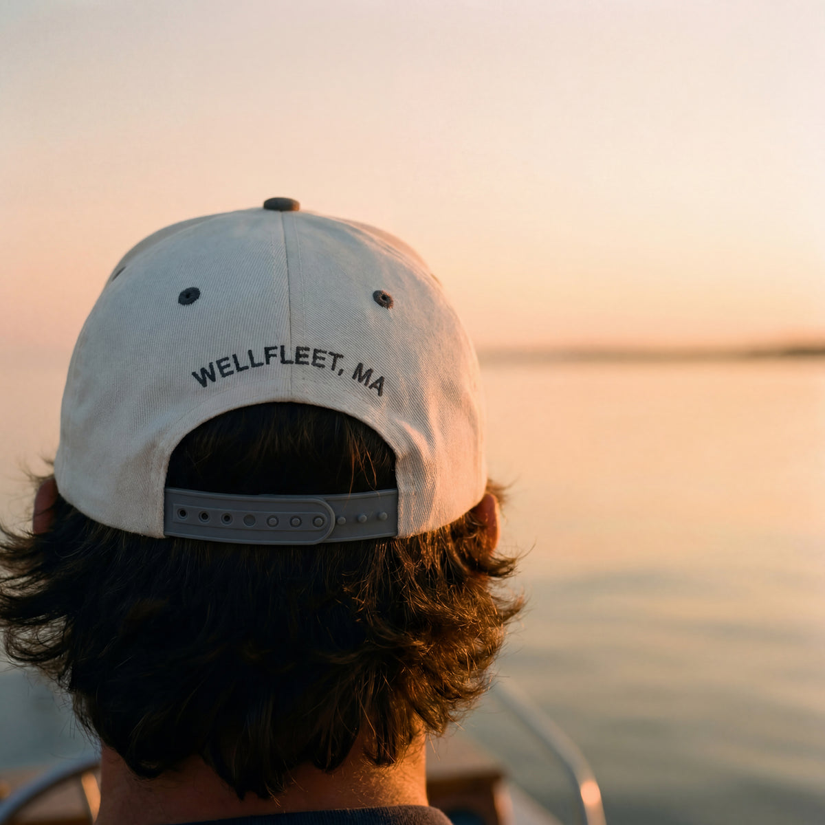 Person wearing a cap with 'Wellfleet, MA' text overlooking a sunset over water.