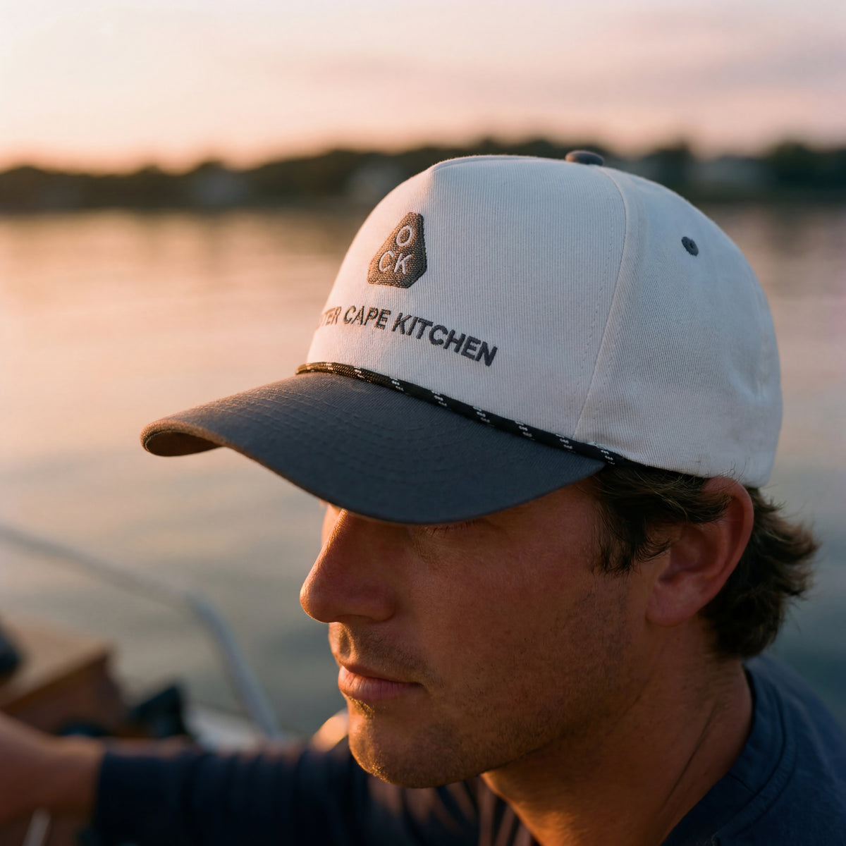 Man wearing a cap with 'Outer Cape Kitchen' logo by a body of water at sunset.